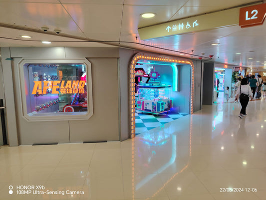 購物中心內一間明亮的、霓虹燈閃爍的電子遊戲室。左側的玻璃顯示器上有「Apeland 屯灣廣場」的標誌。閃亮的地板反射著燈光。人們走過。品牌:工程案例,顯示日期:2024/08/22。.