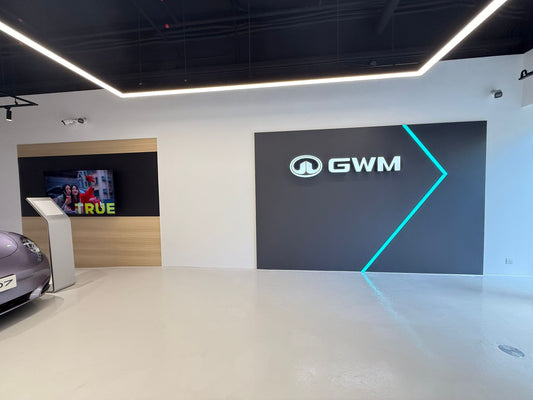 展示室的牆上掛滿了工程案例的標誌和品牌。前景中可以看到 Ora 車行展廳的汽車引擎蓋,而電視螢幕左側則顯示有「TRUE」字樣的影像。.