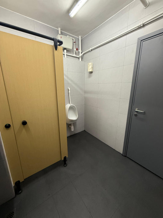 由工程案例(牛奶公司貨倉碼頭廁所裝修)設計的公共廁所設有米色隔間門、小便池、灰色瓷磚牆壁和門,配有高架螢光燈和可見管道。.